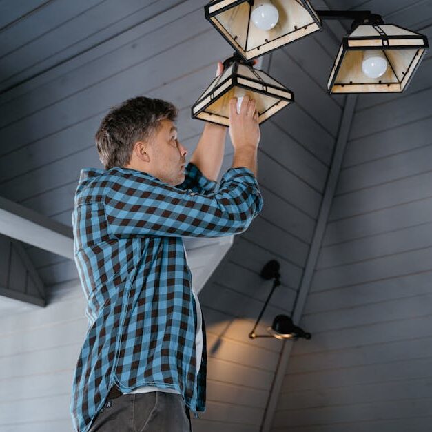 A man in a plaid shirt changes a light bulb on a ceiling lamp in a stylish, modern interior.
